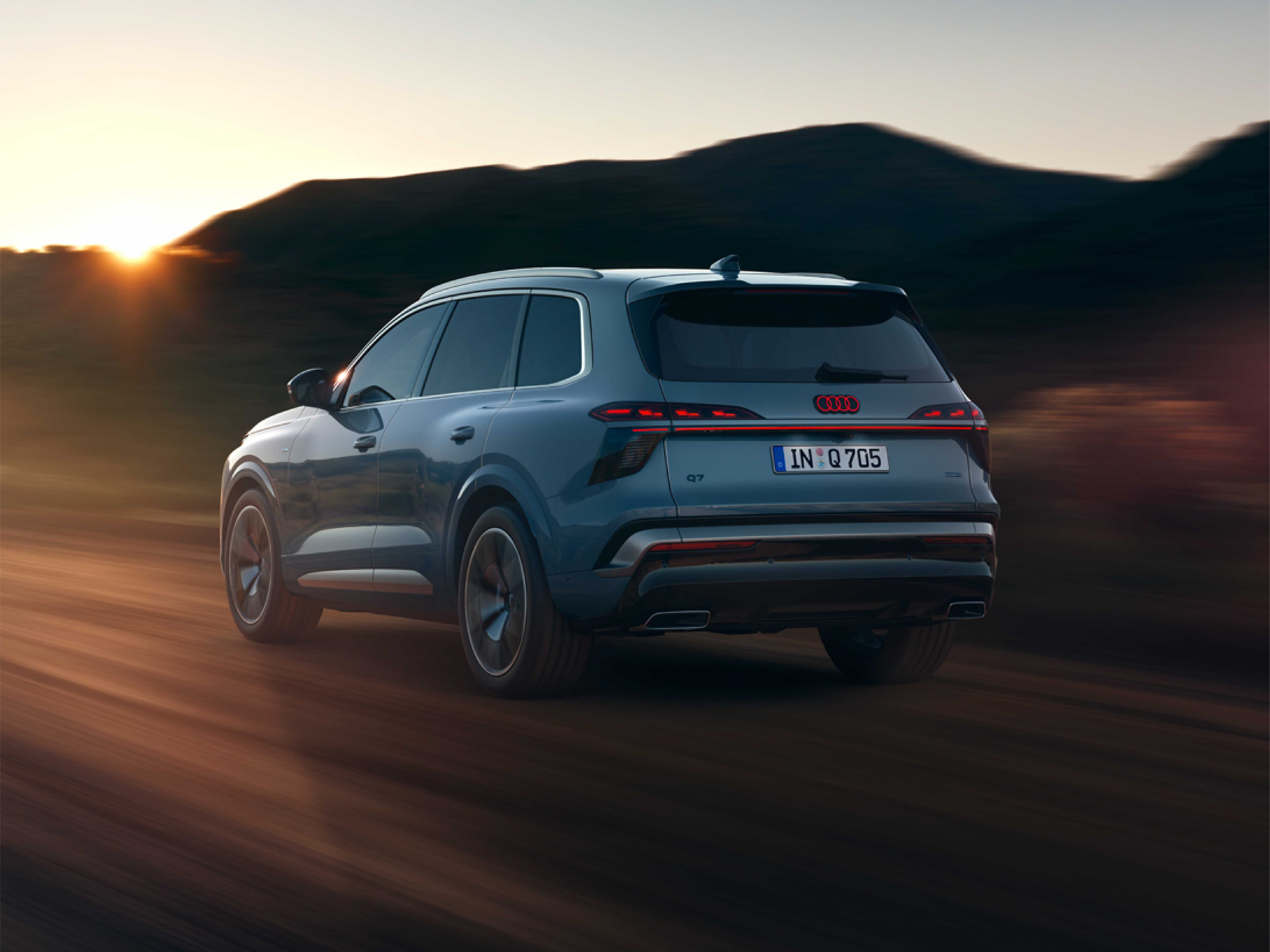 Alt-Text: Heckansicht eines graublauen Audi Q7, der dynamisch auf einer unbefestigten Straße fährt. Zu sehen sind das rote LED-Rücklichtband, die Modellbezeichnung „Q7“ und das Kennzeichen „IN Q 705“ vor einer Hügellandschaft bei Sonnenuntergang.